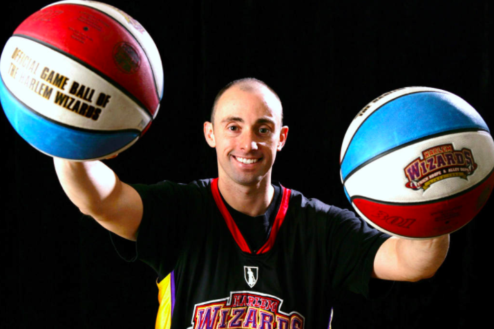Image Description: Mike Simmell holds two basketballs (one in each hands) in front of a black backdrop. He smiles for the camera.