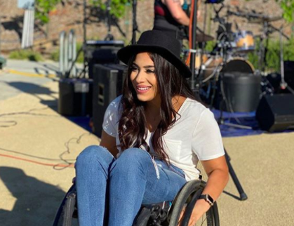 Image Description: Steph Aiello sits in her wheel chair, popping a wheelie. She smiles gleefully.